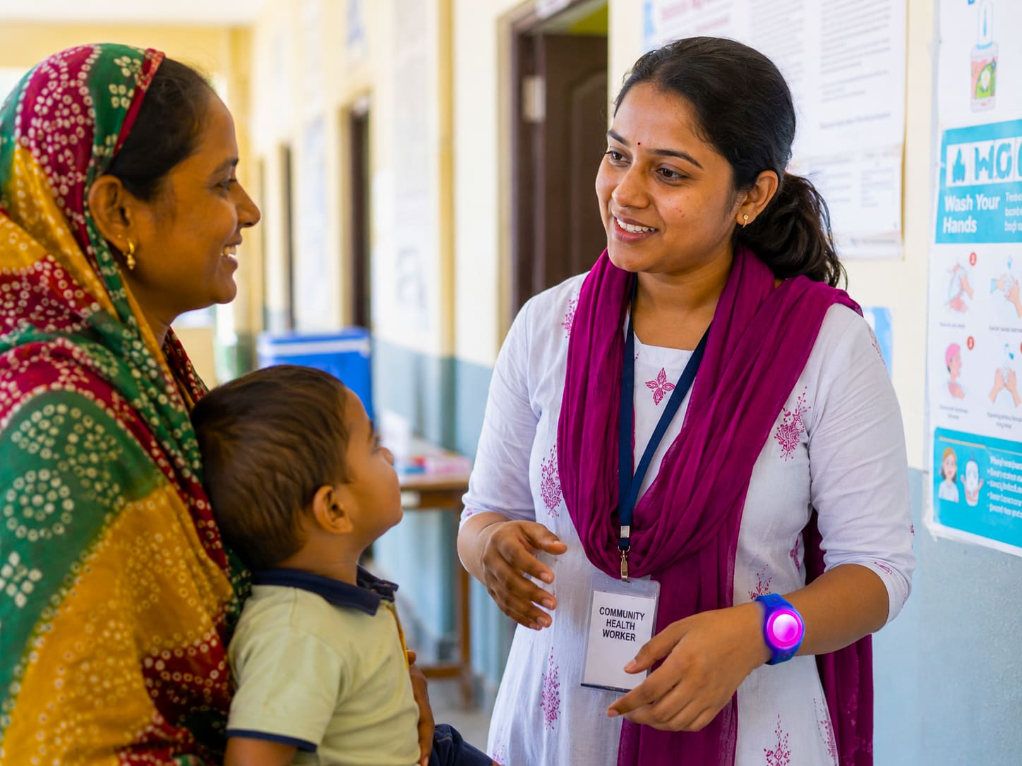 A community health worker in India speaking with a mother and child, wearing a magenta WearSpray band on her wrist
